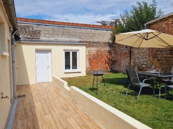 A private outdoor space is featured, showcasing a grassy area bordered by light-colored walls. A patio set with a table and chairs is positioned under a large umbrella, while a charcoal grill stands nearby. Natural light brightens the scene.