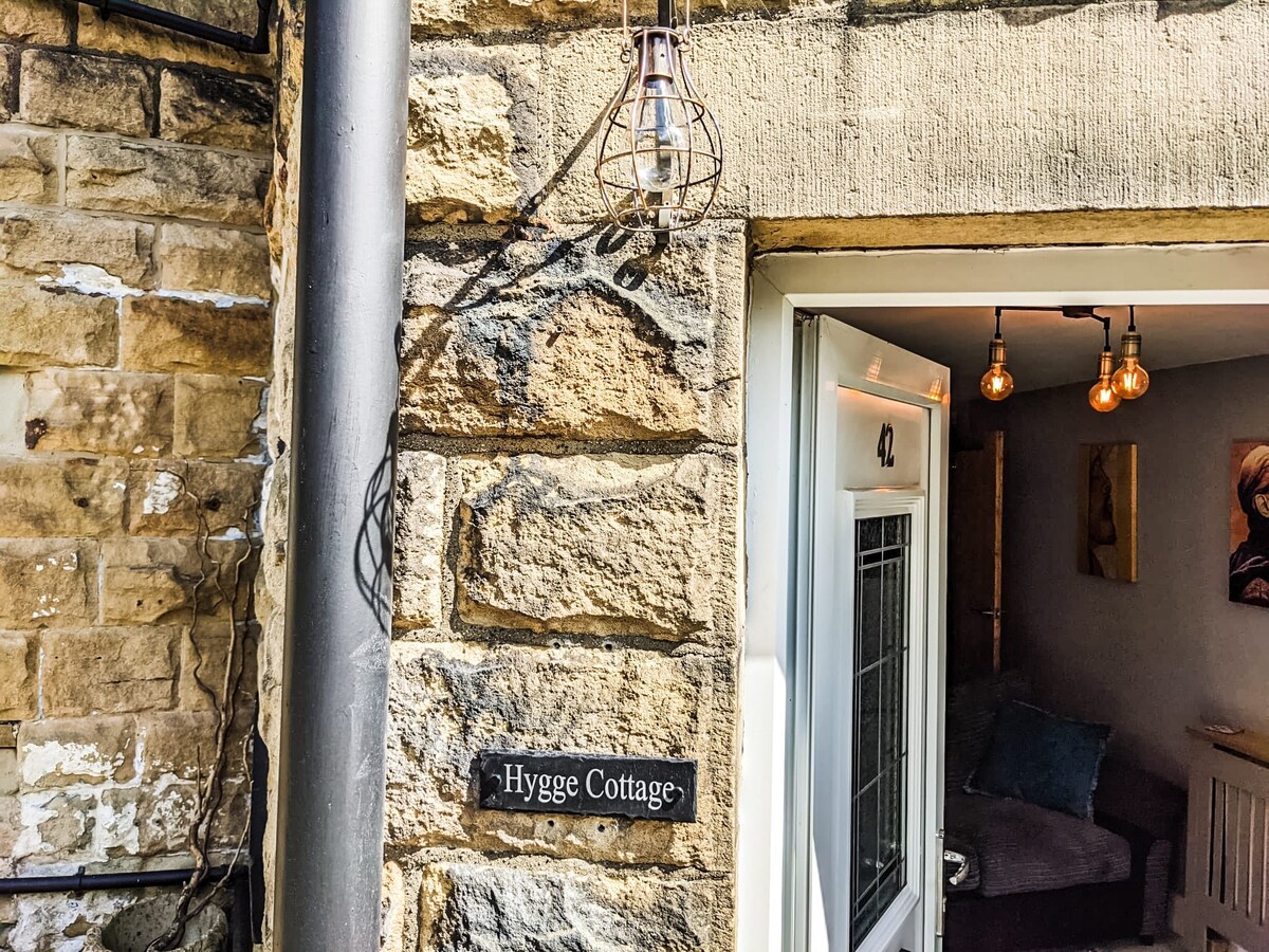 The entrance to Hygge Cottage is framed by natural stone walls. A sign displays the name of the cottage, while warm light bulbs hang above the entrance. The door opens to reveal a glimpse of the cozy interior, inviting exploration.