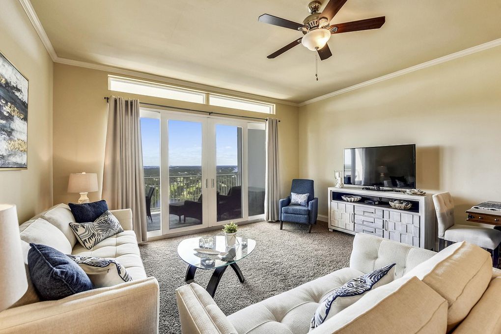 A bright living room is furnished with two comfortable sofas and accent chairs. A glass coffee table sits in the center, and a large television is positioned against the wall. Expansive glass doors provide views of the outdoors and bring in natural light.