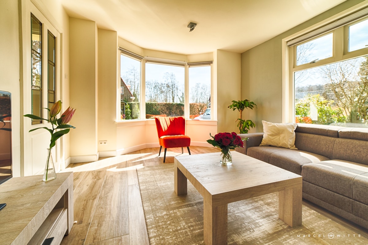 A sunlit living area is presented with a modern sofa in neutral tones and a light wood coffee table. A vibrant red chair adds a pop of color, while a flower arrangement and a potted plant provide natural accents. Large windows offer views of the outdoor greenery.