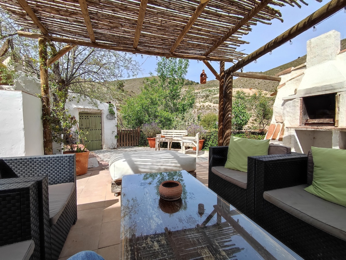 An outdoor terrace features a seating area with dark wicker furniture and light green cushions. A wooden dining table is visible in the background, surrounded by greenery. An outdoor grill is situated beside the seating area, enhancing the inviting atmosphere of the space.