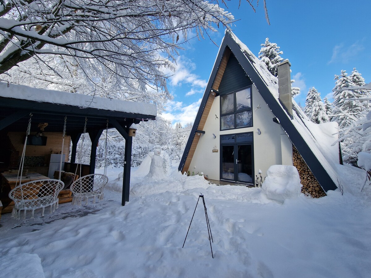 An A-frame cabin is partially covered in snow, with large windows reflecting the blue sky. Outdoor seating is provided, featuring white chairs. A nearby stack of firewood is visible. Trees, blanketed in snow, frame the space, enhancing the serene atmosphere.