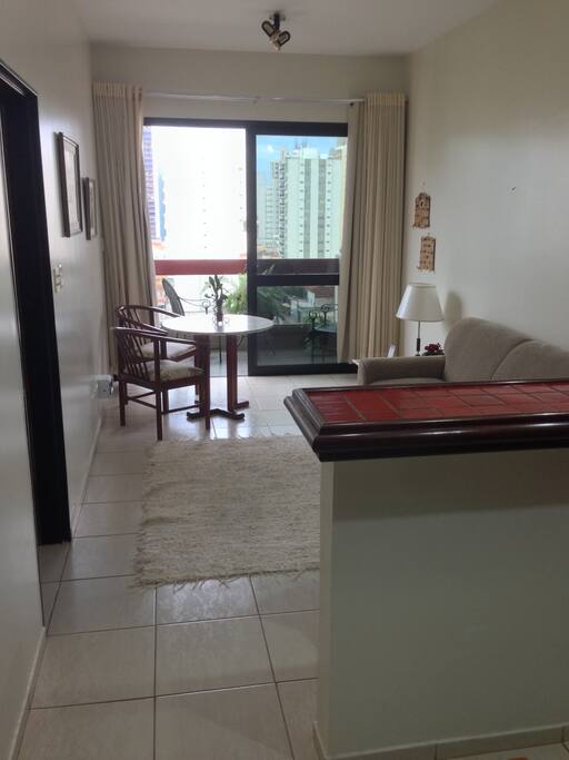 Sala com mesa, duas cadeiras, sofá dois lugares, mesinha de apoio com abajur e telefone, ventilador de teto com controle remoto que também controla a luz, cortina dupla, sendo uma blackout.