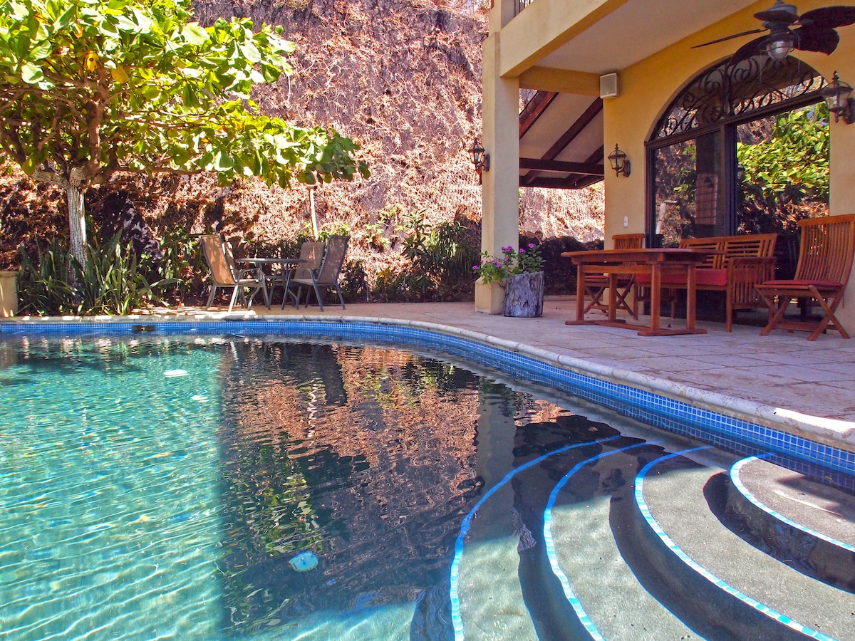 A private swimming pool is featured with gently curved steps leading into the water. Outdoor seating is available, with a wooden dining table and chairs positioned under a shaded overhang. Lush greenery surrounds the area, enhancing the sense of seclusion.