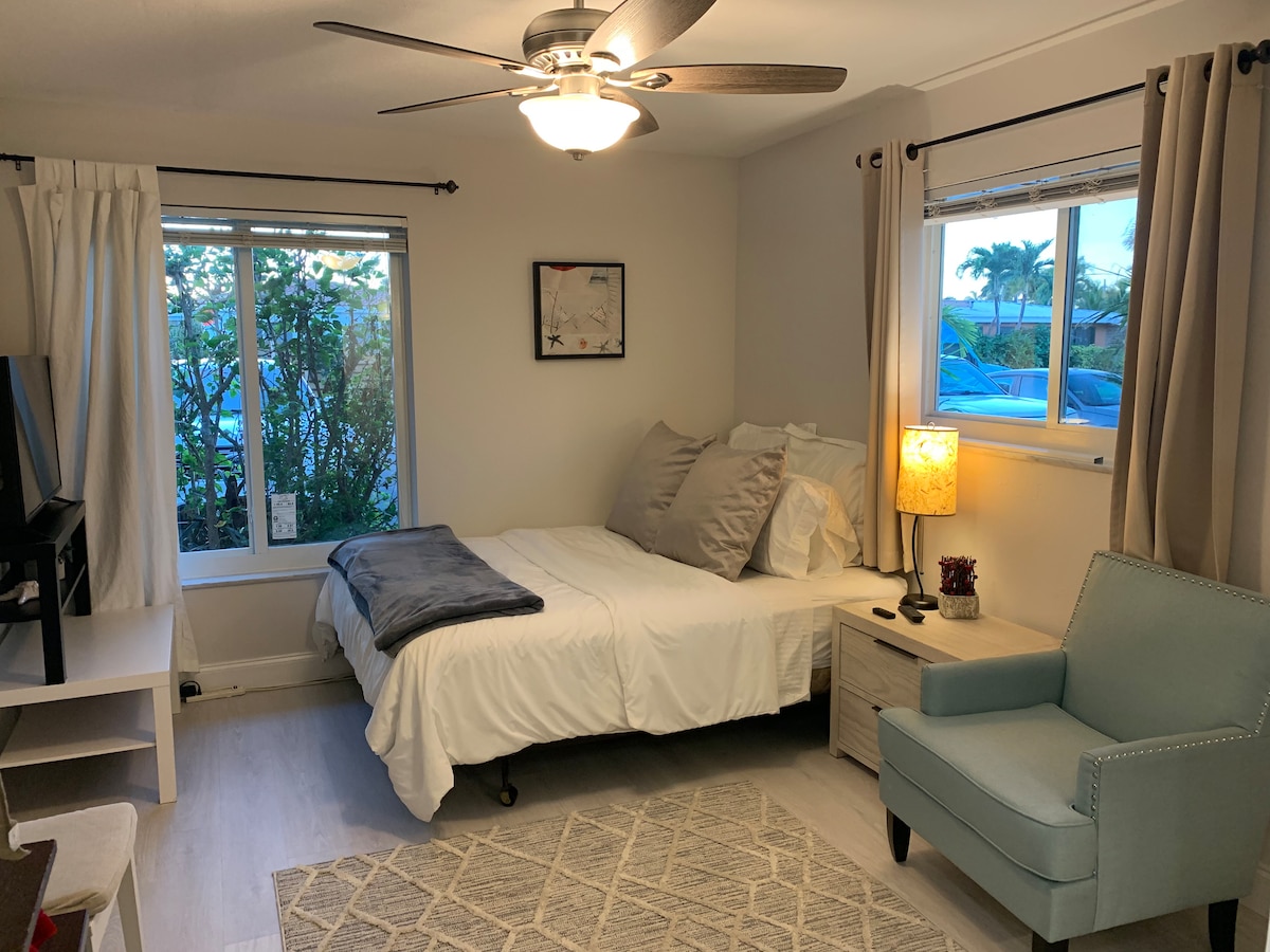 A cozy studio space features a spacious bed adorned with plush pillows and a grey throw. A small nightstand hosts a lamp, while a light blue armchair adds a pop of color. Two windows allow soft natural light to brighten the room, revealing greenery outside.