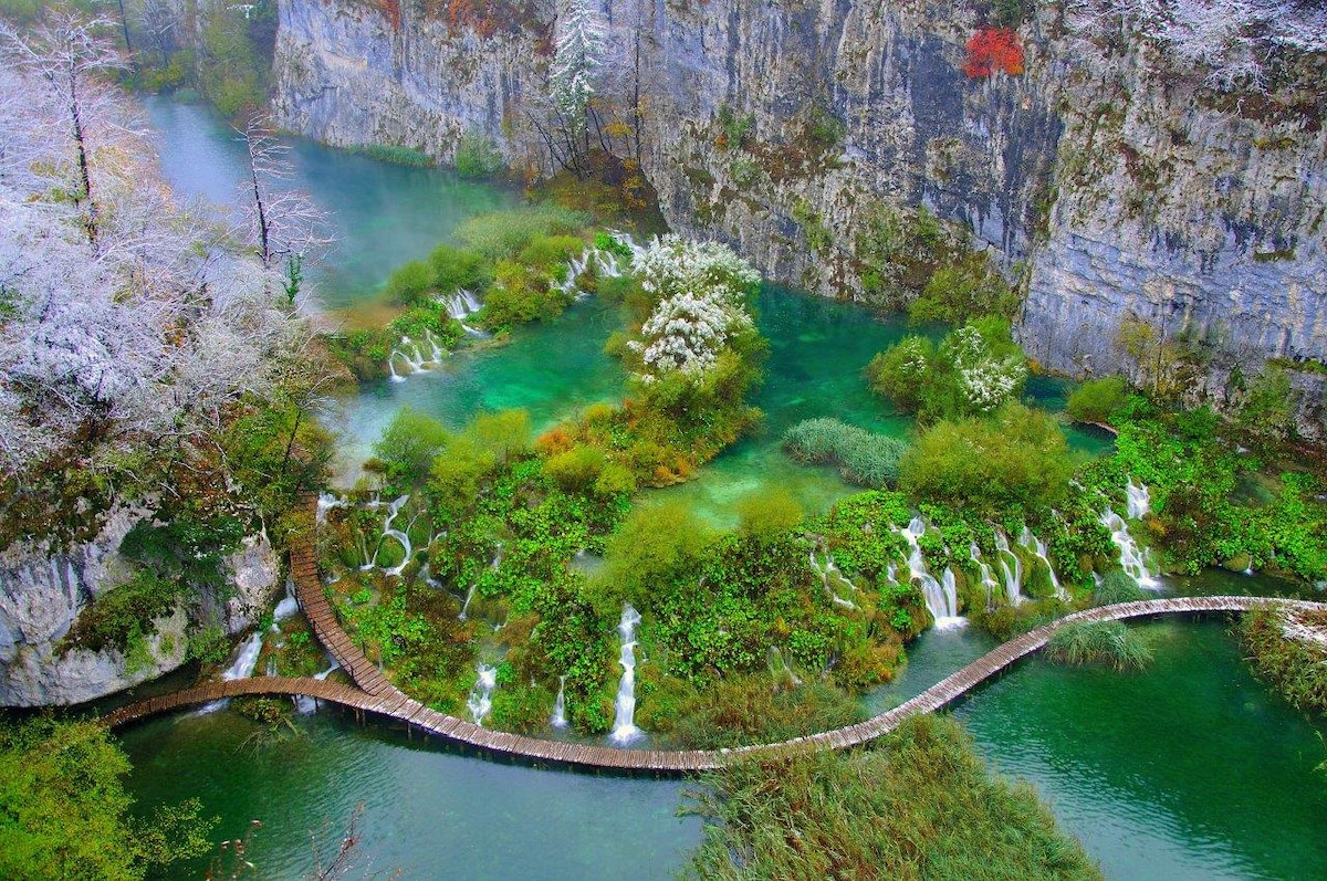 A winding wooden path traverses lush greenery and tranquil turquoise water. Cascading waterfalls are visible throughout the scene, surrounded by rocky cliffs and patches of blooming flowers. The landscape showcases a harmonious blend of nature's colors and textures, inviting exploration and reflection.