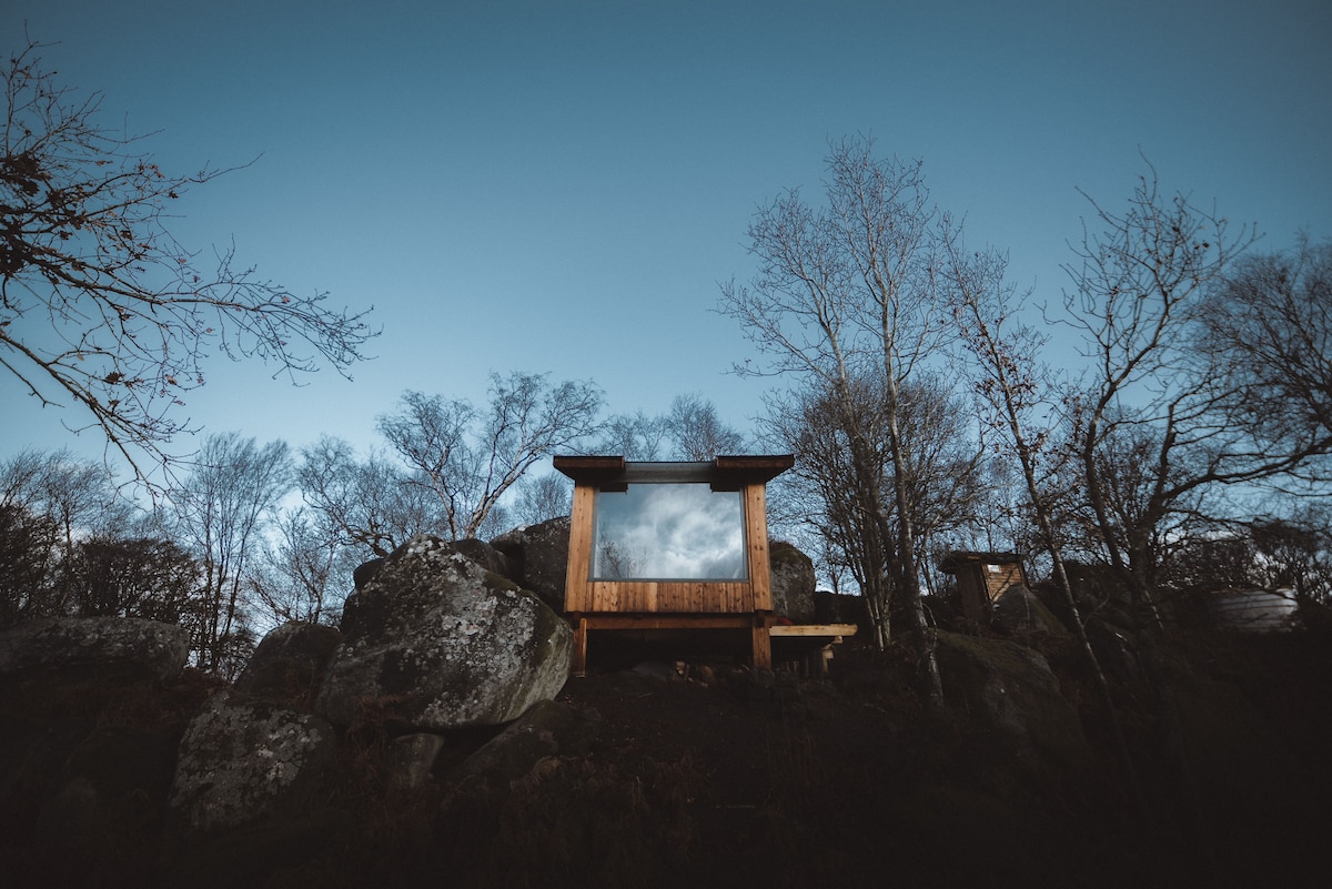 A rustic cabin is situated among large boulders and trees, showcasing large windows that reflect the sky. The natural surroundings are highlighted by a clear blue backdrop, creating a serene atmosphere of blending with nature.