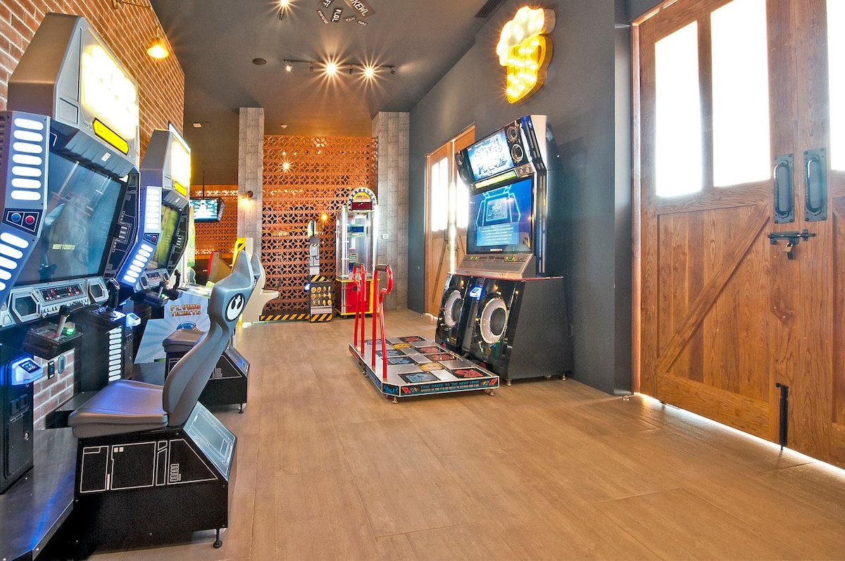 A game room is featured with several arcade machines, including racing and dance games. Natural light enters through large wooden doors, highlighting the spacious layout. The flooring is composed of light-colored tiles, enhancing the playful environment of the gaming area.