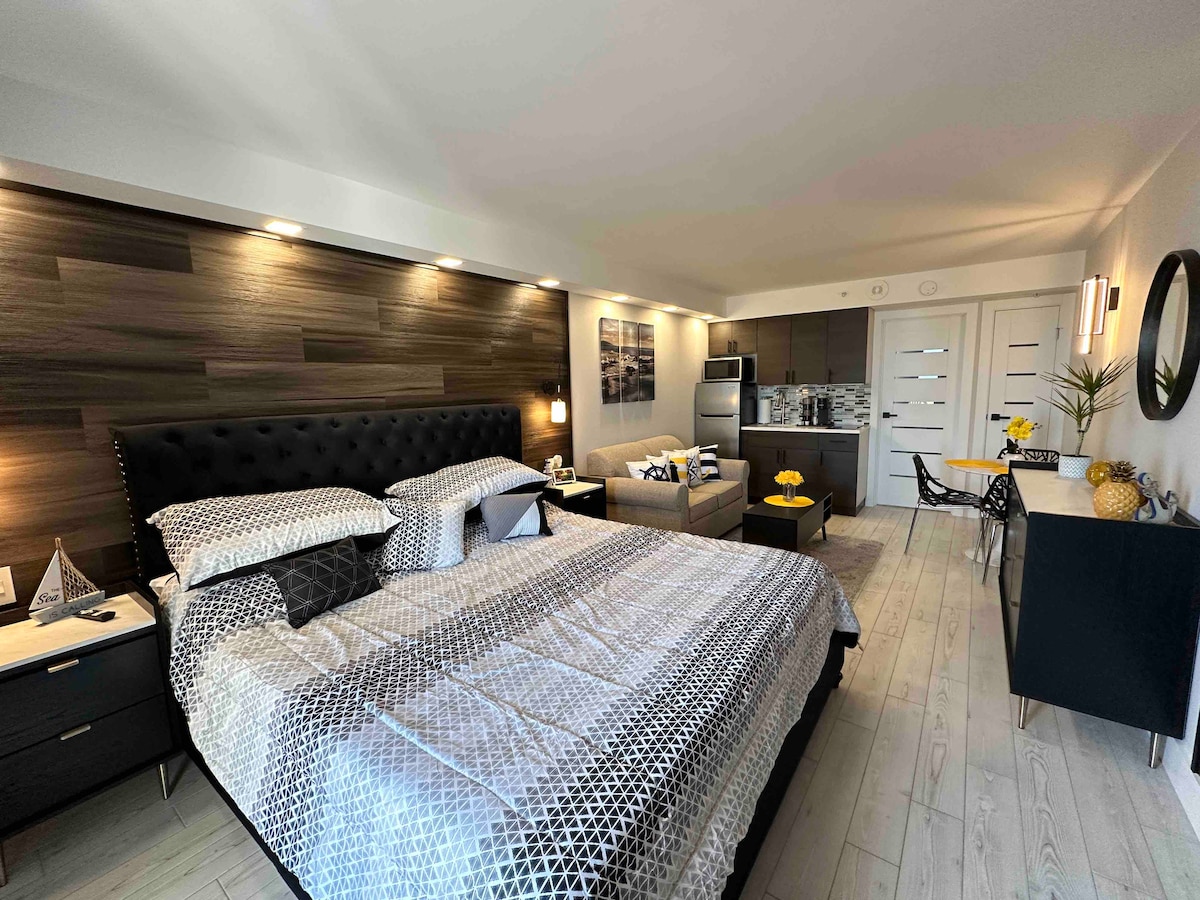 A spacious studio is presented with a king bed adorned in a geometric patterned duvet. Modern cabinetry and stainless steel appliances are visible in the kitchen area. A cozy seating space features a sofa and small dining table, blending comfort and functionality.