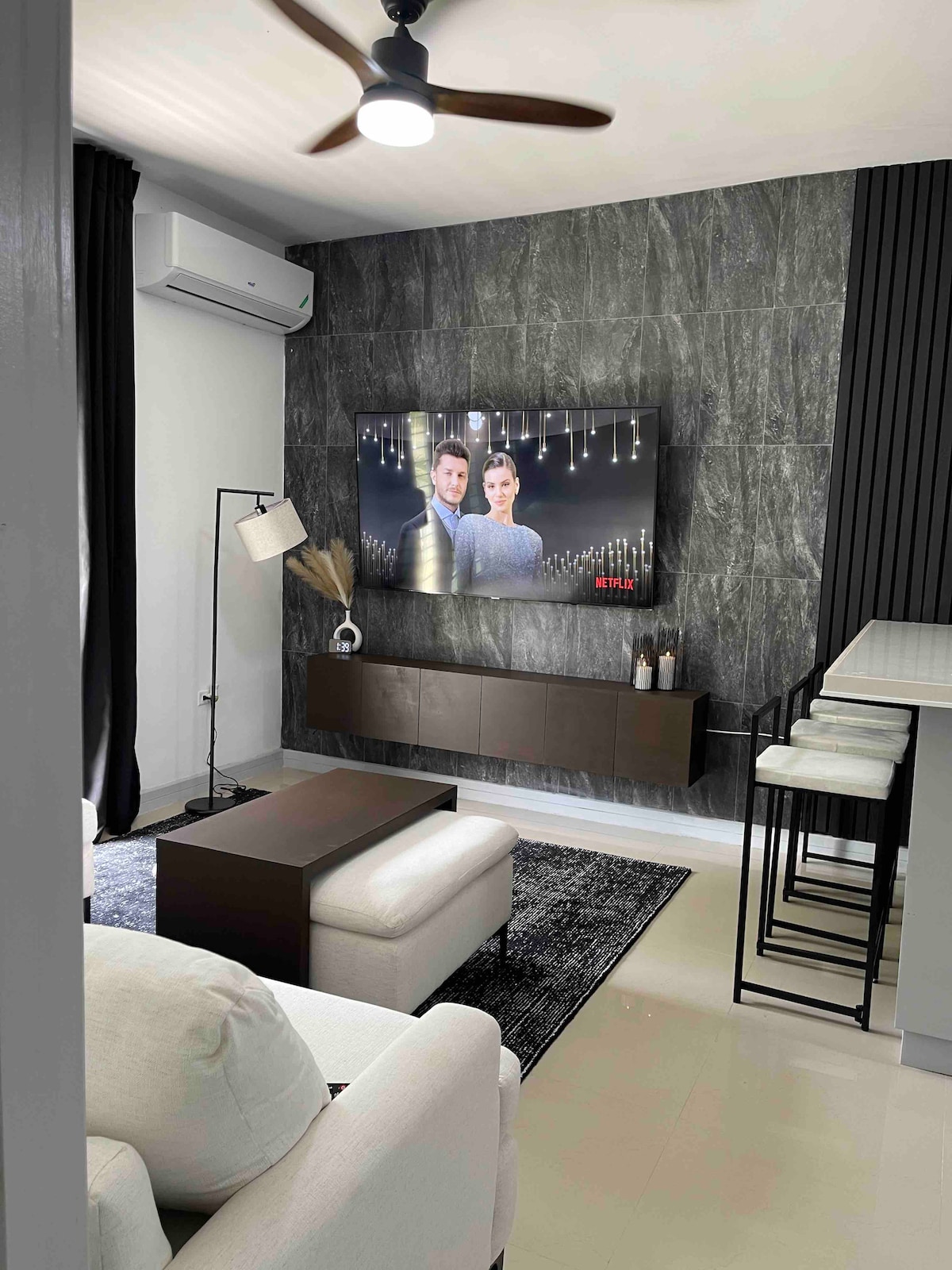 A modern living area features a sleek wall-mounted television against a textured gray backdrop. A comfortable white seating arrangement is positioned in front, accompanied by a stylish floor lamp. The open space includes a high table with bar stools visible in the background.