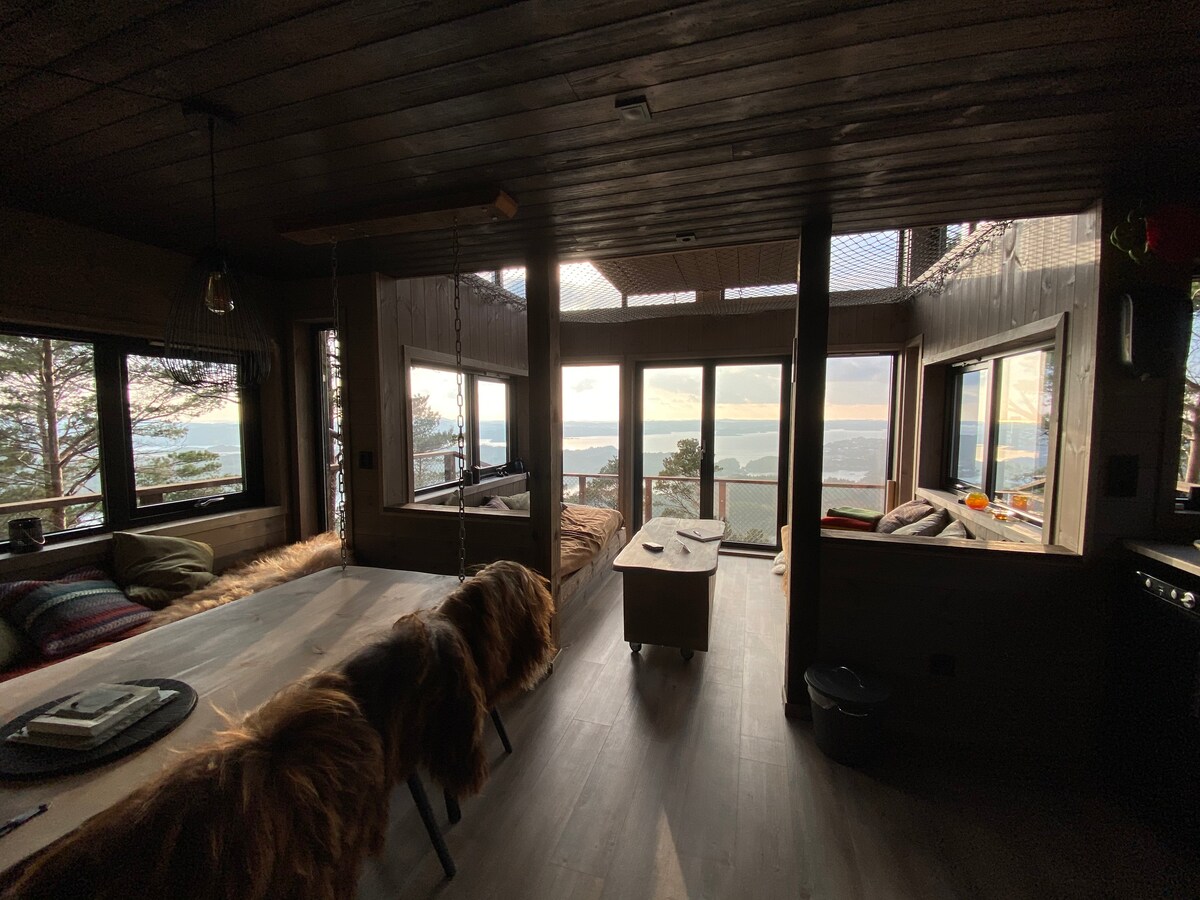 A living area is seen featuring large windows that frame a scenic view of the landscape. Natural light floods the space, illuminating a wooden dining table and cozy seating. Textured rugs and warm furnishings add a touch of comfort to the atmosphere.