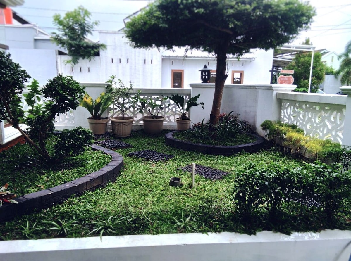 An indoor garden is featured, characterized by lush greenery and various plants arranged in decorative pots. A circular pathway composed of dark stones winds through the garden, bordered by trimmed hedges and flowering plants, creating a serene and inviting space.