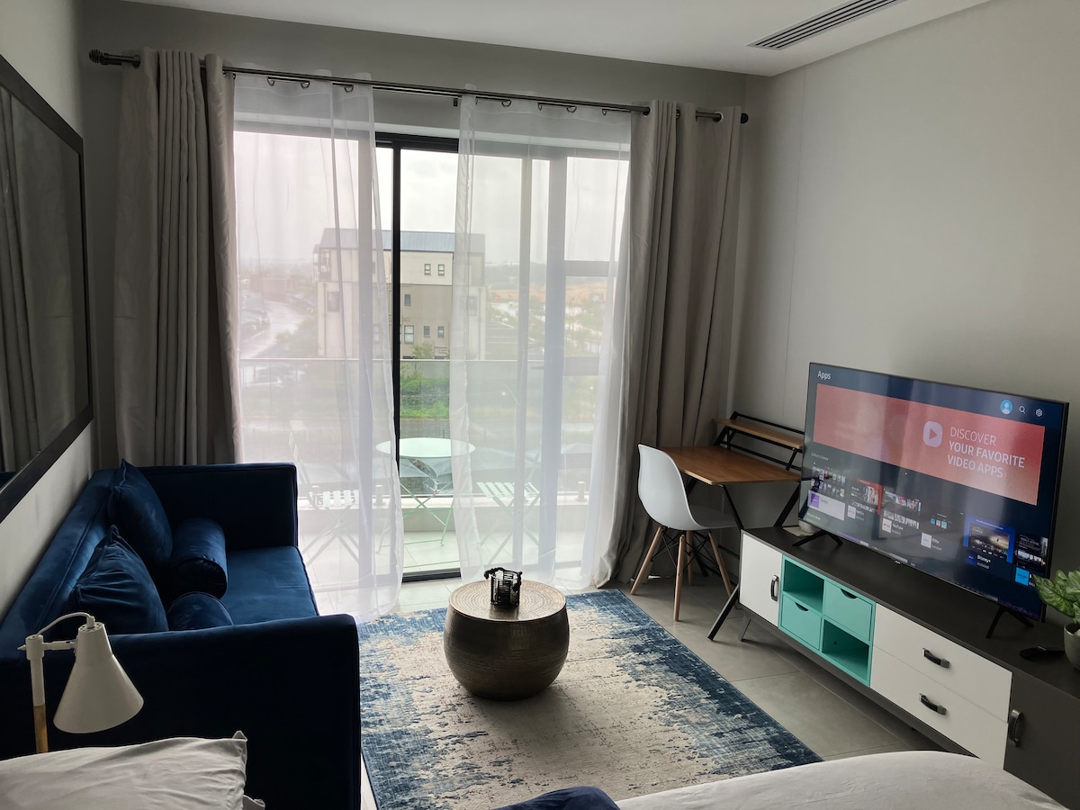 A spacious living area features a blue sofa and a round coffee table, complemented by a light rug. A television stands on a low console, while a workstation with a chair is positioned near the large glass sliding door, allowing natural light to this comfortable space.