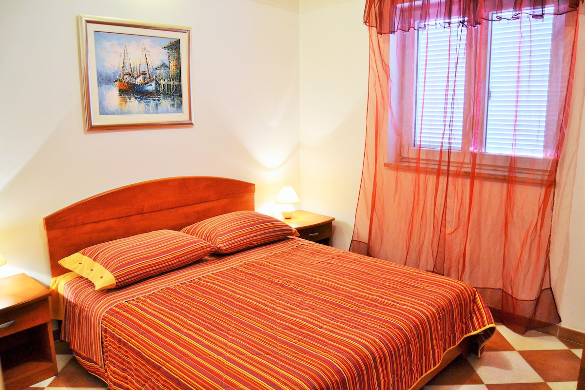A sleeping area is presented with a wooden bed featuring a striped red and yellow bedspread. Two bedside tables are positioned alongside the bed. Soft lighting is provided by two lamps, and a colorful painting adorns the wall. Natural light enters through a window covered with sheer curtains.