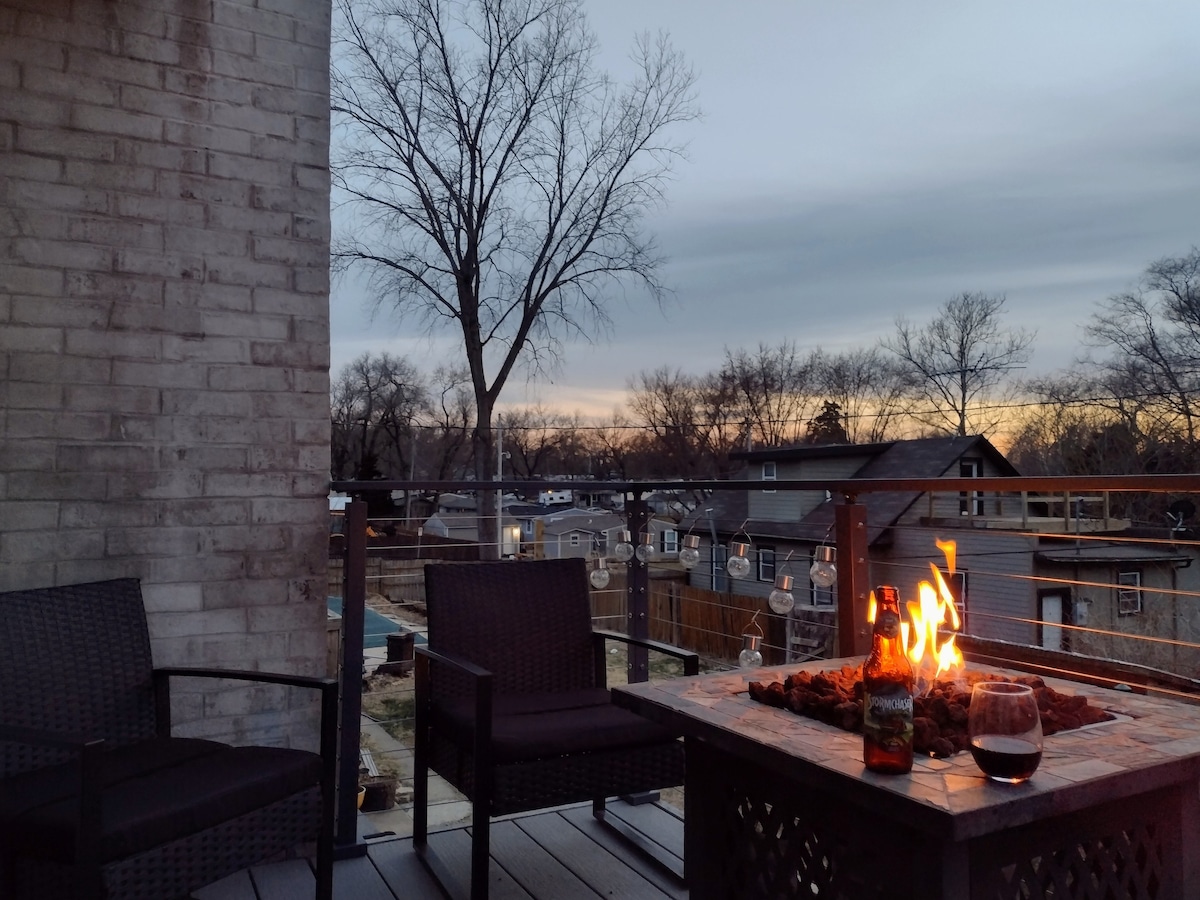 A peaceful outdoor space features a fire pit surrounded by wicker chairs, with a drink resting on the table. In the background, a sunset casts a warm glow over neighboring homes and trees, creating a serene atmosphere.