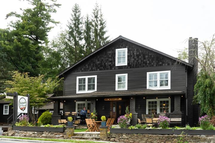 The exterior of the inn features a two-story black wood building with large windows and a welcoming entrance. Comfortable seating arrangements with chairs are placed on a stone patio surrounded by vibrant greenery and blooming flowers, inviting guests to relax outdoors.
