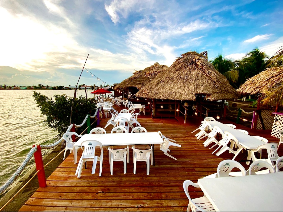 A spacious wooden deck overlooks the water, furnished with several white plastic tables and chairs. Thatched roof structures are seen in the background, accompanied by palm trees and a clear blue sky. The setting invites relaxation by the water.