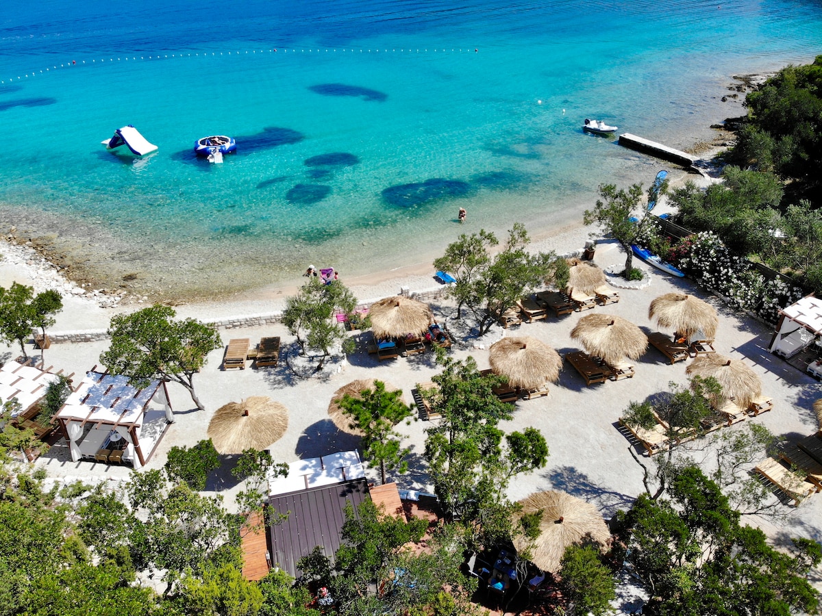 A sandy beach is visible with crystal clear blue waters gently lapping at the shore. Several straw huts provide shaded seating areas along the beach, surrounded by greenery. Small boats can be seen in the water, alongside floating platforms, contributing to a serene coastal setting.