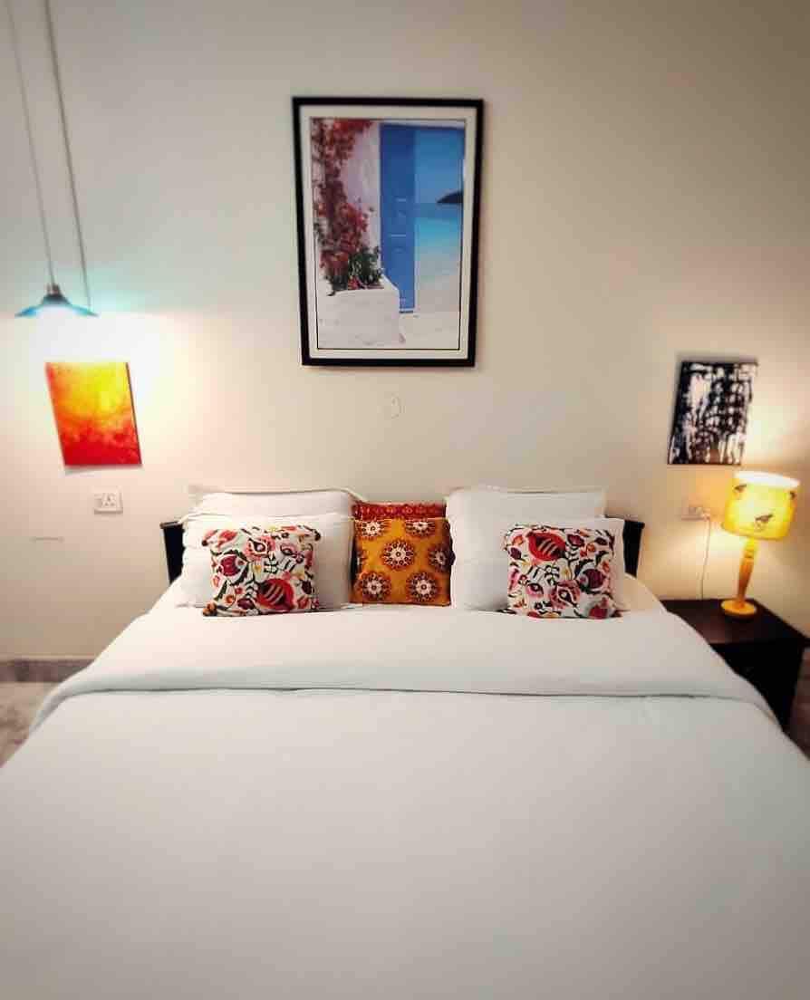 A cozy bedroom features a spacious bed adorned with decorative cushions and a soft white blanket. A bedside lamp emits a warm glow, while vibrant artwork decorates the walls, providing a touch of color. Natural light is welcomed through a nearby window.