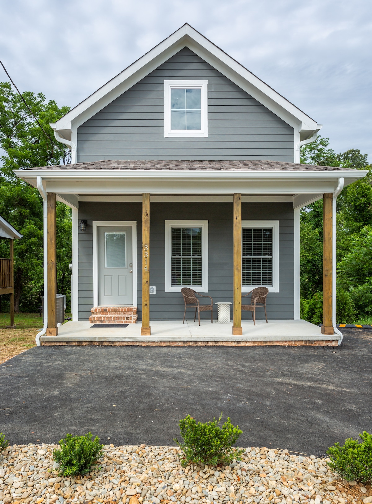 A two-story gray cabin features a welcoming front porch with two chairs and a small table. The front door is framed by large windows, offering glimpses of the natural surroundings. Gravel and landscaping adorn the entrance, enhancing the outdoor space.