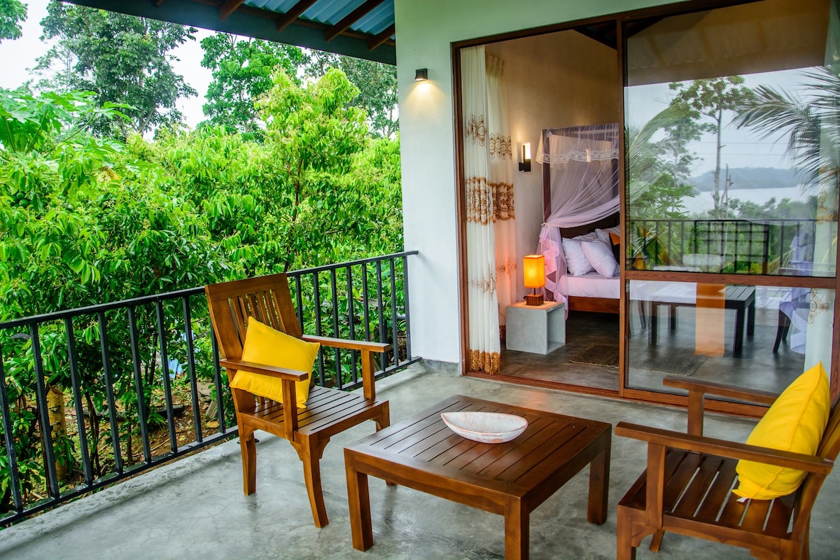 A covered veranda features two wooden armchairs and a small table, offering a view of lush greenery and Beratuduwa Lake. A large sliding glass door connects to a well-lit room, where a spacious bed is visible through sheer curtains.