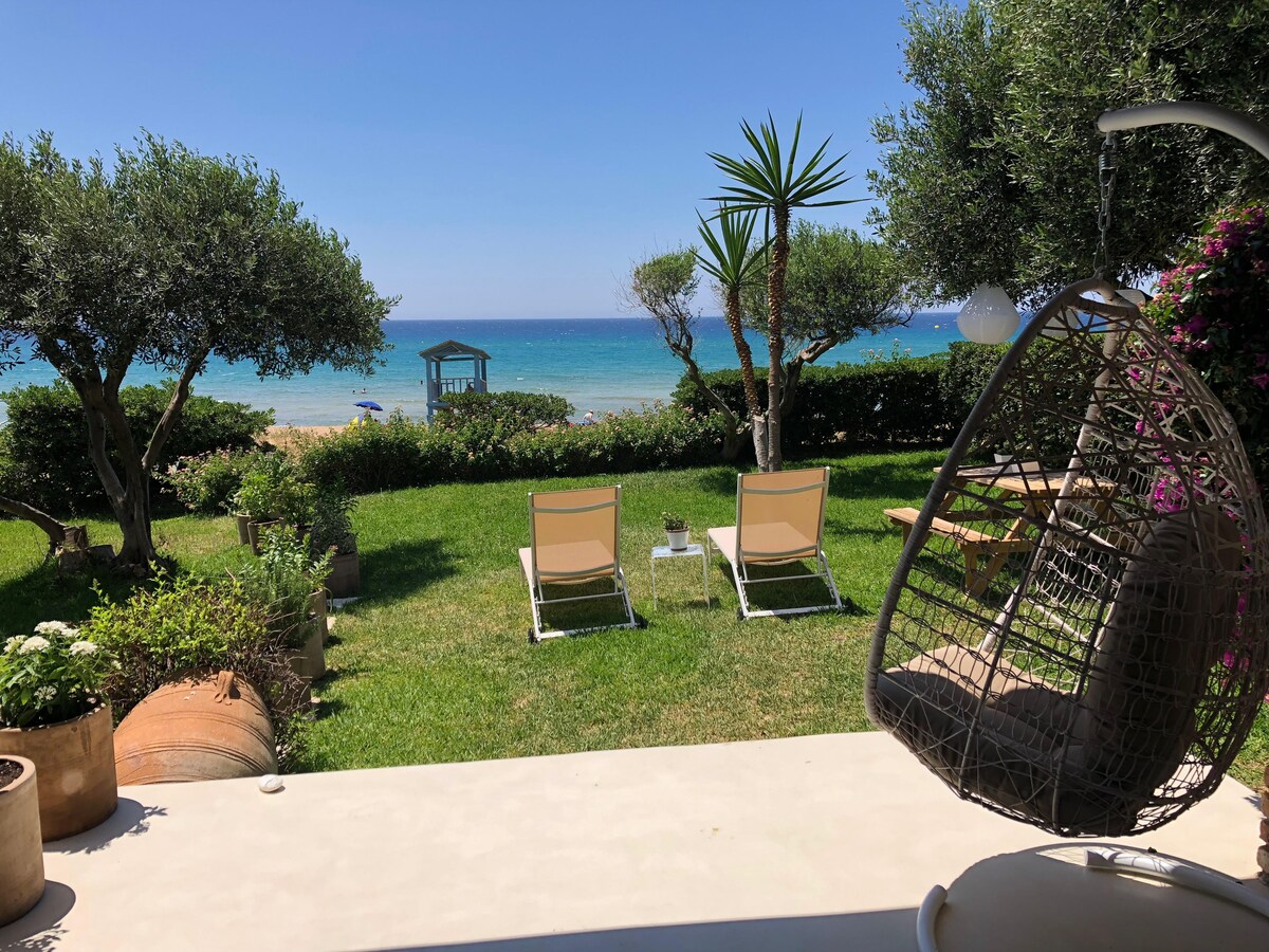 A private garden area features two lounge chairs positioned on well-maintained grass, overlooking the beach. Lush greenery, including olive trees and palm plants, frames the view of the calm blue sea. A small gazebo is visible in the distance, enhancing the peaceful atmosphere.