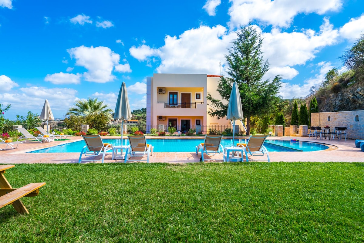 A large swimming pool is surrounded by sun loungers and umbrellas, set against a backdrop of lush greenery and clear blue skies. The villa is visible in the background, showcasing a spacious structure with balconies. Invitations to relax are offered by the well-kept garden and pool area.