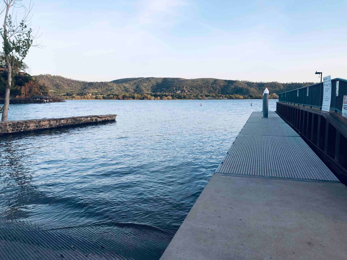 A tranquil lake view is captured, showcasing a smooth surface reflecting the sky. A wooden dock extends into the water, with gentle waves lapping at its edge. Rolling hills can be seen in the background, adding depth to the serene landscape.
