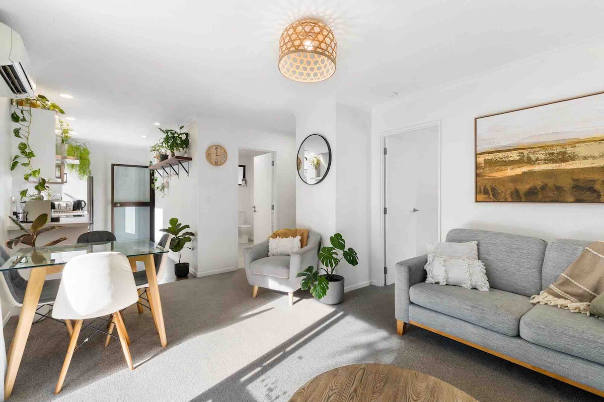 The spacious living area features natural light illuminating a comfortable grey sofa and a small armchair. A modern dining table with white chairs is visible, alongside a decorative wall clock and plant accents. A doorway leads to the kitchen and a separate area in the background.