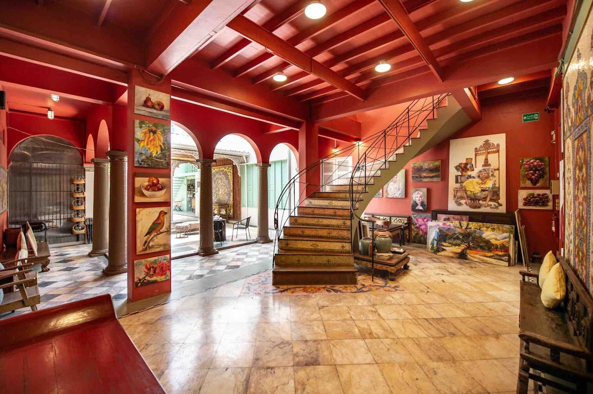 A vibrant grand foyer features a striking red color scheme, complemented by ornate staircases and arched windows. Artwork lines the walls, showcasing various styles and subjects. Marble flooring adds elegance, and seating areas can be seen in a cozy, inviting arrangement.