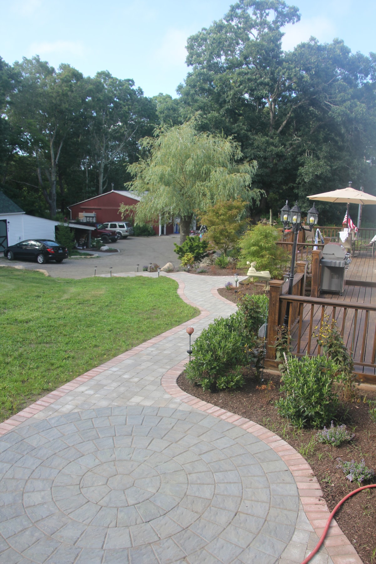 A stone pathway winds through a well-maintained yard, leading towards a wooden deck. Lush greenery and decorative plants are visible along the path, while a spacious parking area and a red barn-like structure are seen in the background, surrounded by trees.