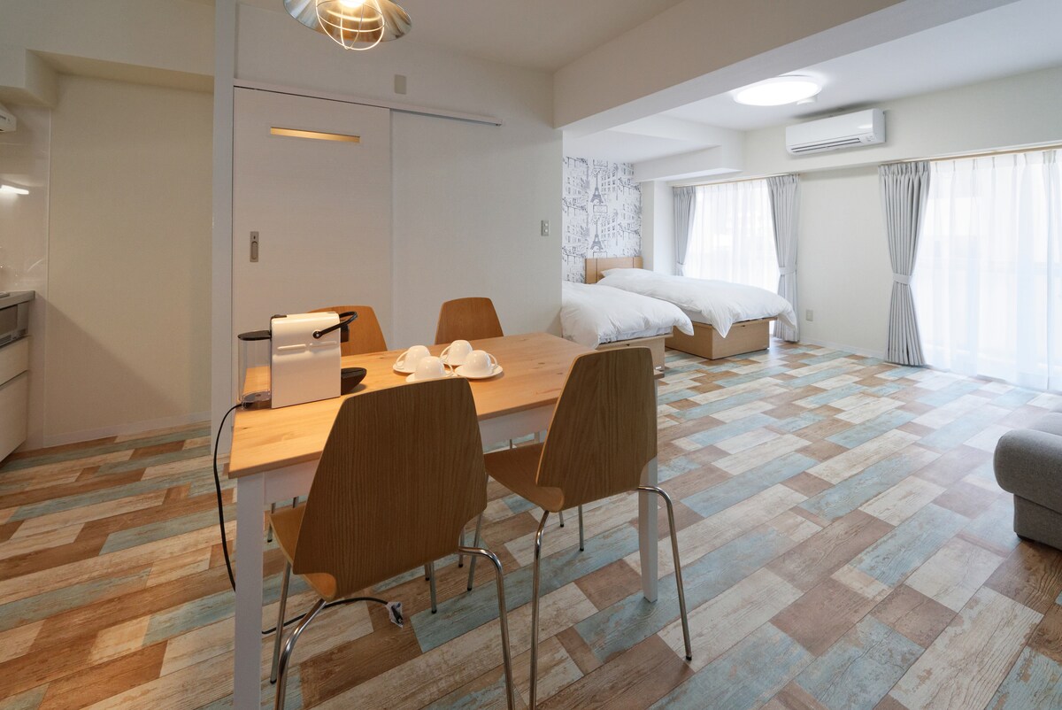 A spacious room features a wooden dining table with four chairs, and a comfortable bed situated near large windows draped with light curtains. The floor showcases a mix of natural wood tones, contributing to a bright and airy environment.
