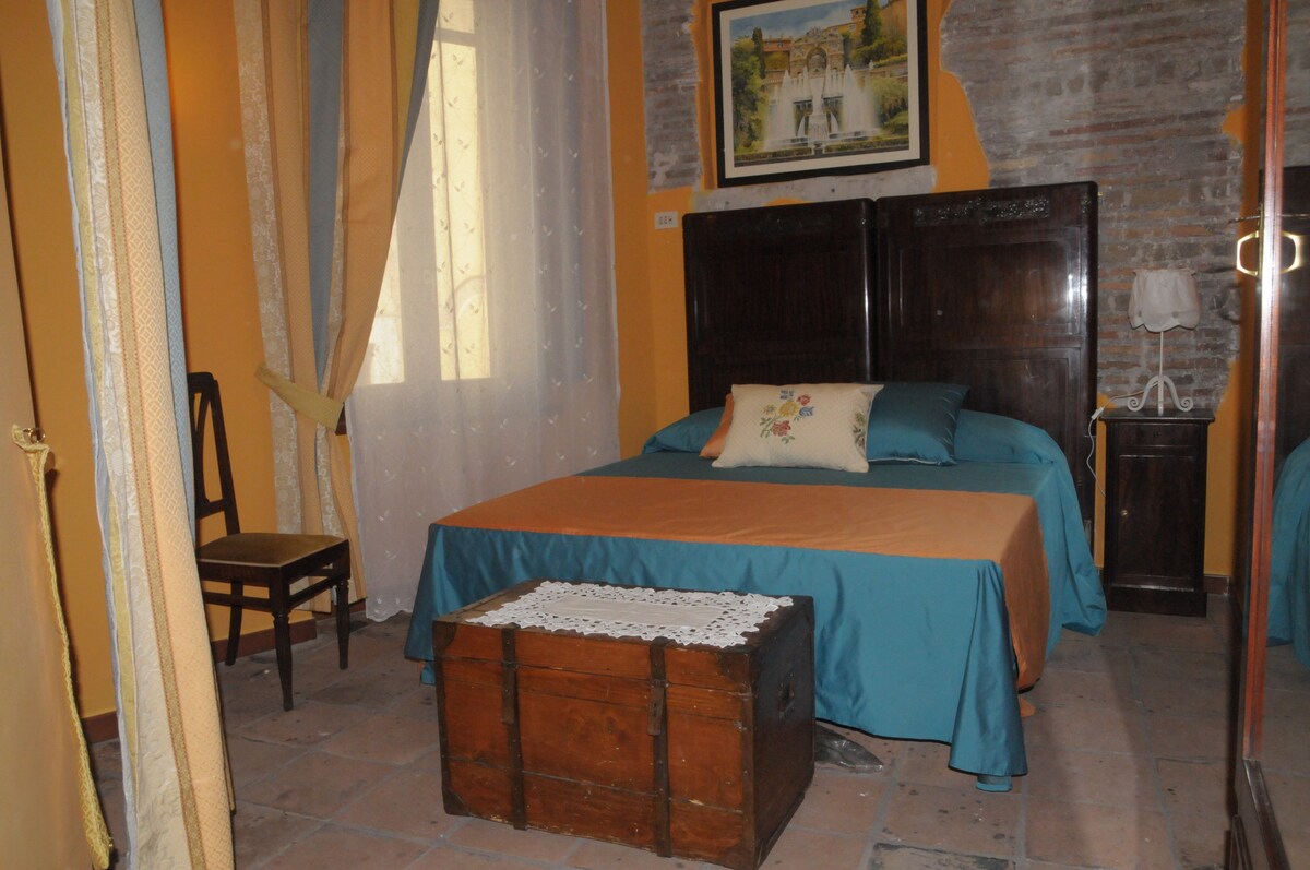 A serene bedroom is presented with a spacious bed adorned in light blue and orange linens. A rustic wooden trunk is positioned at the foot of the bed, complementing the warm-colored walls. A chair and window draped with delicate curtains are also visible.