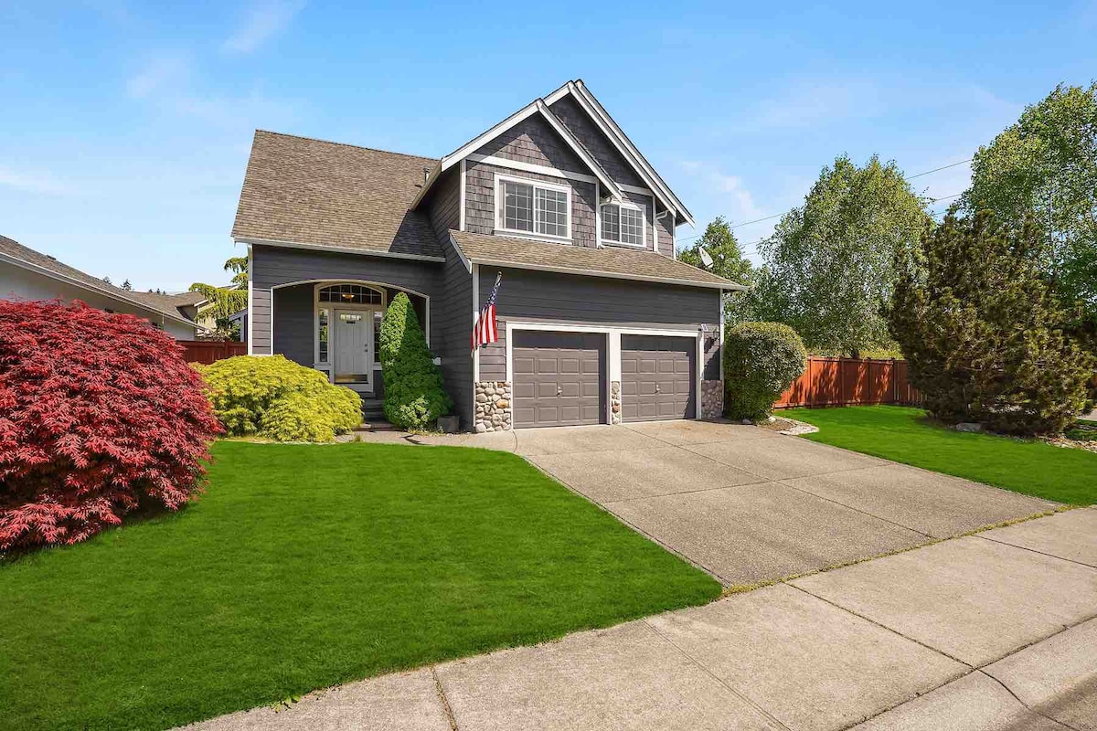 A two-story home is depicted with a well-maintained exterior. The front lawn features lush green grass and colorful shrubs, complemented by a welcoming pathway leading to the front door. A driveway accommodates two vehicles, and an American flag proudly hangs from the porch.
