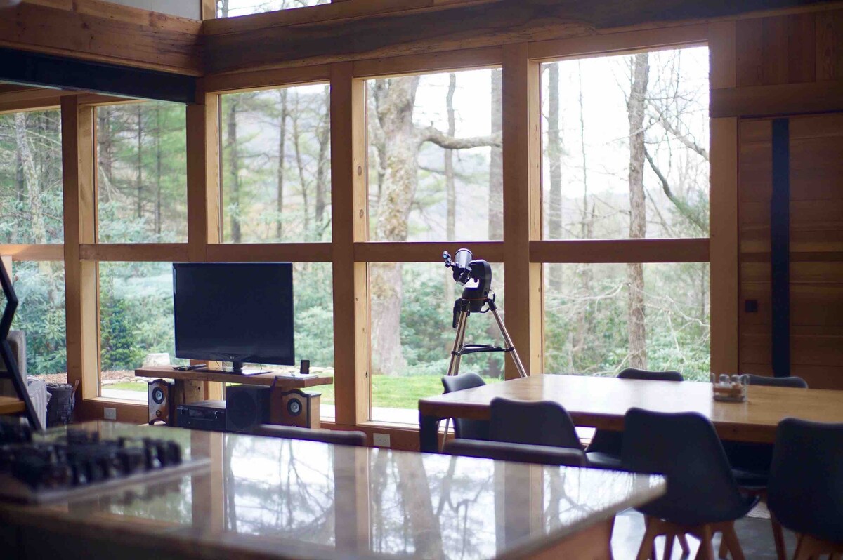 A modern living area features large windows that invite natural light and provide views of the surrounding trees. A television is positioned across from a dining table. A telescope is set near the window, allowing for exploration of the outdoor scenery.