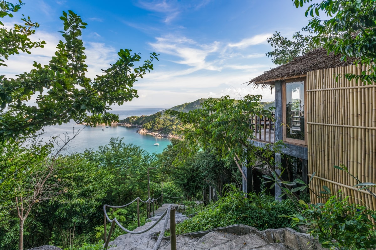A scenic view captures the lush greenery surrounding a bamboo bungalow perched on a hillside. The turquoise waters below are framed by rocky shores and distant hills, creating a serene atmosphere. A winding stone pathway leads from the bungalow to the water's edge.