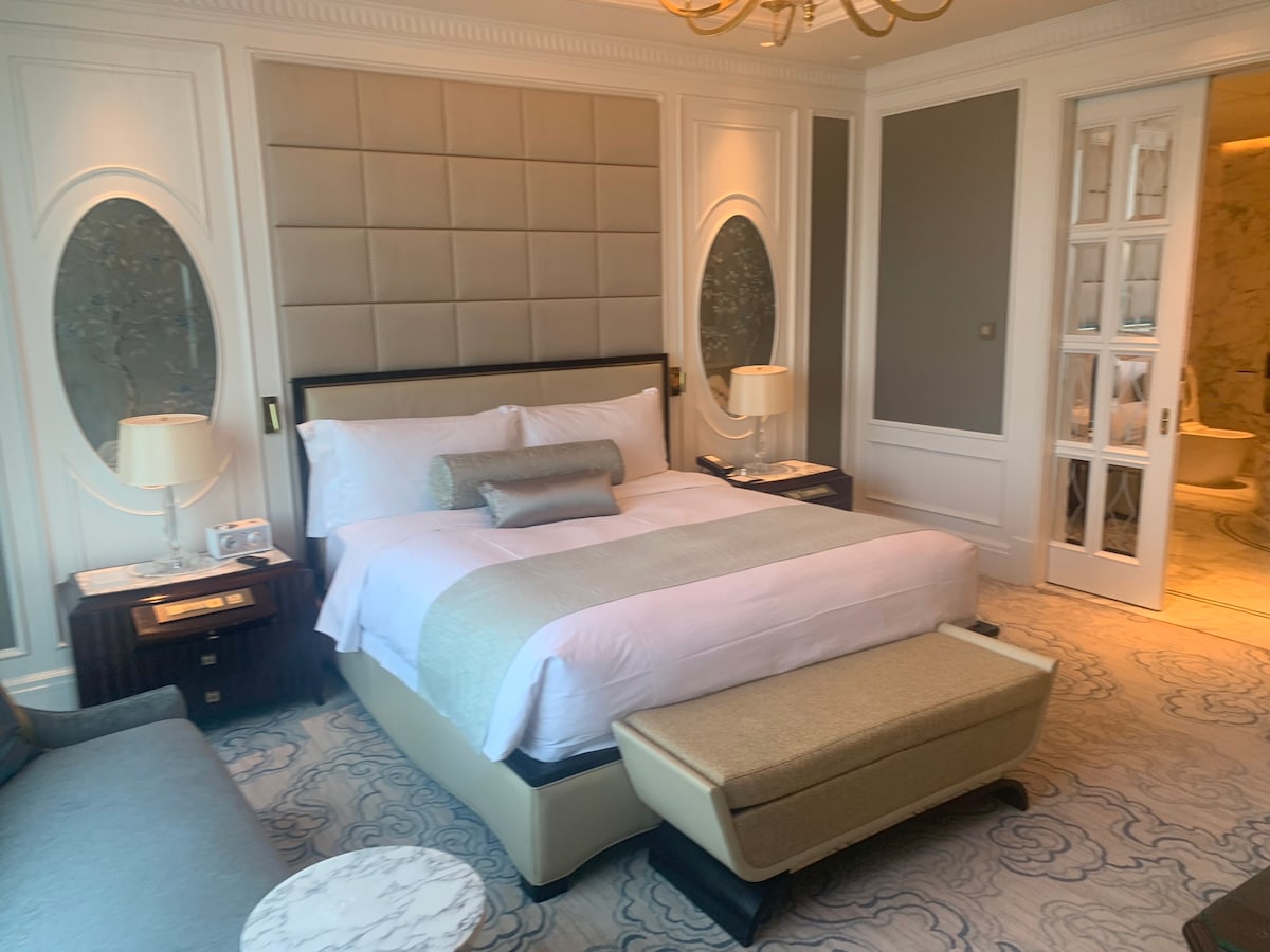 A elegantly furnished hotel room features a spacious bed with soft linens. The neutral color palette is complemented by decorative wall accents and stylish lamps. A cozy bench is positioned at the foot of the bed, while a plush sofa offers additional seating.