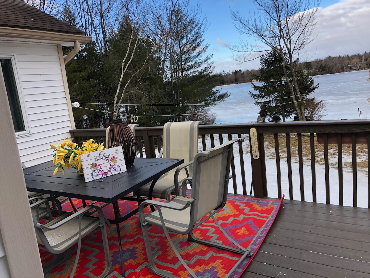 A furnished deck is shown overlooking the serene lake, featuring a dining table surrounded by four chairs. A large area rug offers color beneath the table, while a vase of yellow flowers adds a pop of brightness to the setting. The backdrop includes trees and a frozen lake.