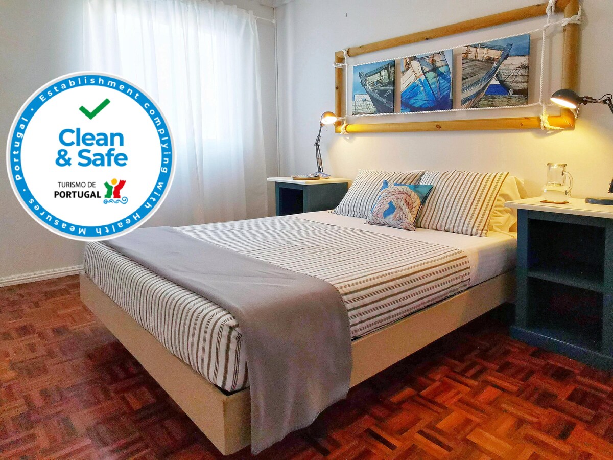 A well-lit sleeping area features a spacious bed with a striped coverlet and decorative pillows. Above the bed, a maritime-themed artwork is displayed within a wooden frame. A blue nightstand holds a reading lamp on either side, with sunlight filtering through a nearby window.