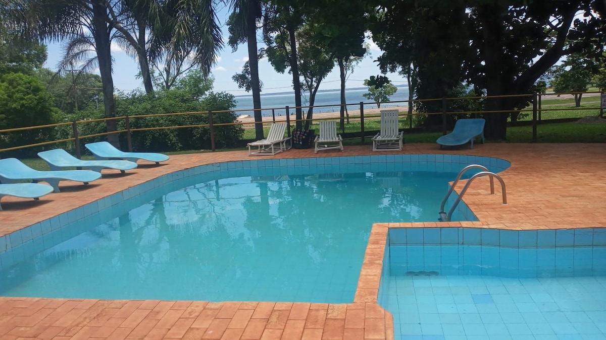 A refreshing swimming pool features clear blue water surrounded by lounge chairs. Lush greenery and palm trees provide shade, while ocean views are visible in the distance. A shallow area near the steps invites relaxation.