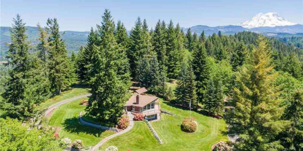 A custom-built cabin is surrounded by lush greenery on a spacious, manicured lawn. Towering coniferous trees frame the property, while the stunning peak of Mt. Rainier is visible in the distance, creating a serene natural backdrop.