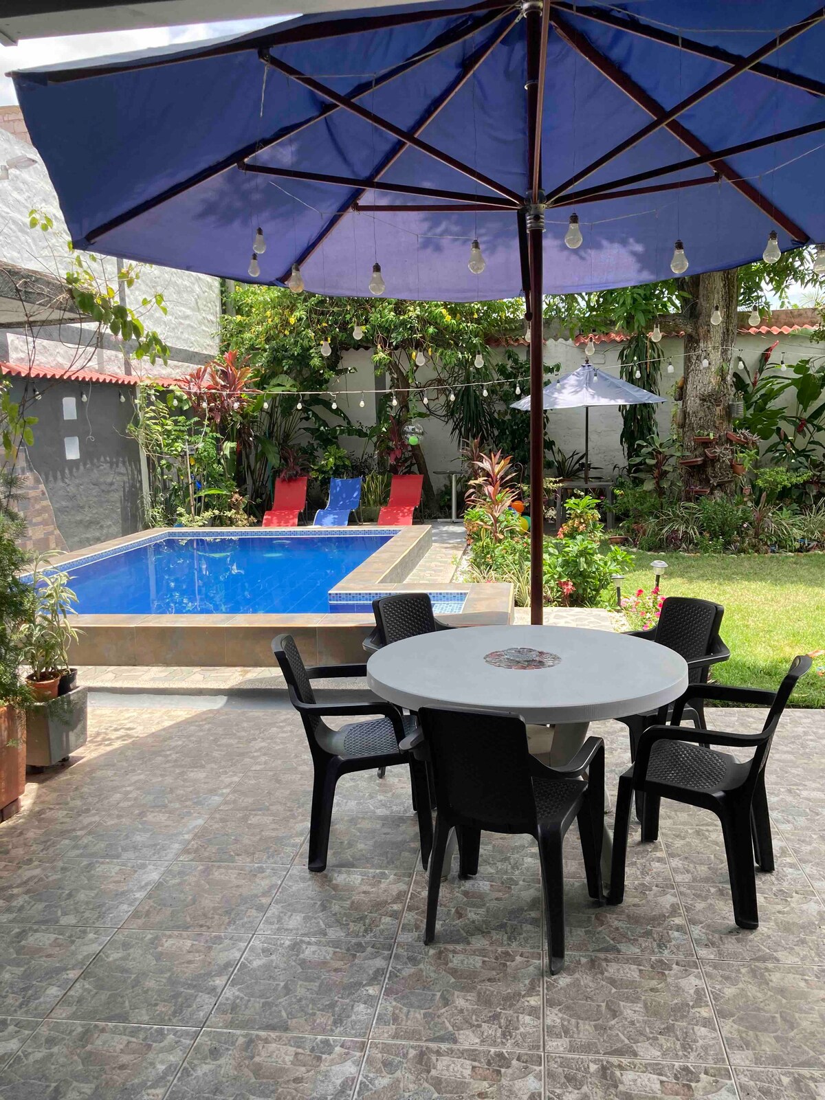 A round table with four chairs is positioned beneath a large umbrella, providing shade in the outdoor area. The well-maintained garden features a refreshing pool nearby, surrounded by vibrant greenery and colorful lounge chairs, creating a relaxing environment.