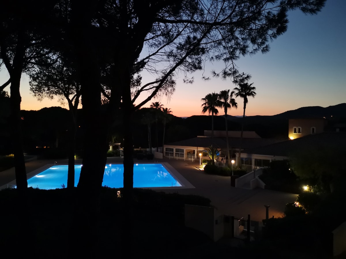 An illuminated swimming pool is visible, reflecting soft blue tones against the evening sky. Surrounding palm trees and bushes create a natural frame, while the distant silhouette of hills adds depth to the landscape. The tranquil ambiance is enhanced by subtle lighting around the pool area.