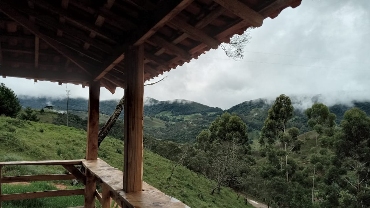 A wooden porch offers a view of rolling green hills and distant mountains, partially shrouded in clouds. The lush landscape is dotted with trees, enhancing the serene rural environment.
