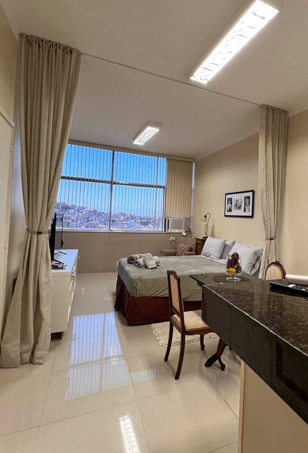 A spacious room features large windows that allow ample natural light. A neatly made bed is visible, accompanied by a small dining table and a chair. Light-colored walls and tile flooring create a bright atmosphere, while decorative elements add a touch of character.