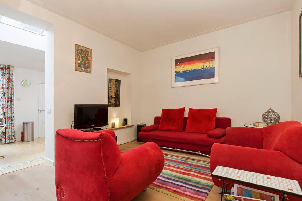 A bright living area features a vibrant red sofa set, complemented by colorful art on the walls. A television is placed on a minimalist stand, and a striped rug adds warmth to the wooden floor. Curtains with playful patterns frame the windows.