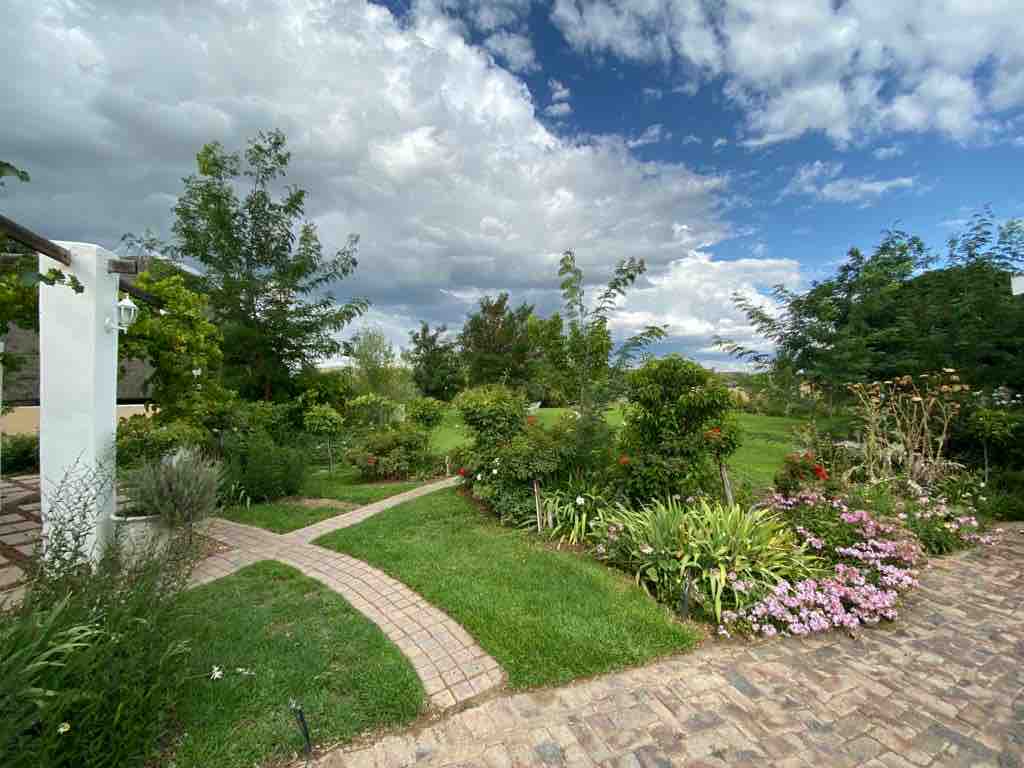 A well-maintained garden area features a winding pathway and diverse plant life, including flowering shrubs and greenery. The sky is partly cloudy, providing a serene backdrop, while a spacious lawn invites relaxation and outdoor activities.