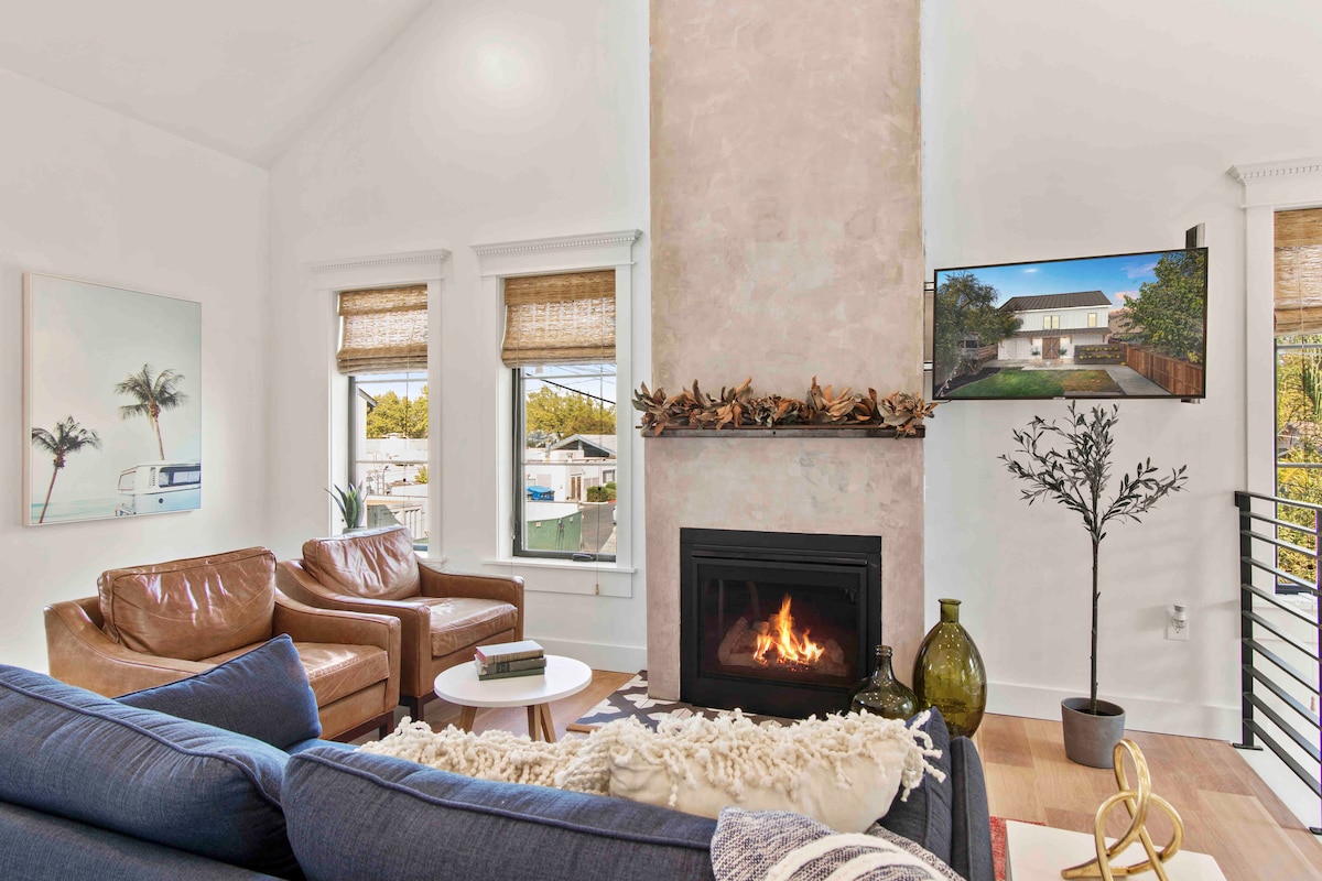 A spacious living room is illuminated by natural light from large windows, featuring a modern fireplace flanked by light-colored walls. A stylish blue sofa is paired with two tan armchairs, while a sleek television is mounted above the fireplace, enhancing the cozy atmosphere.