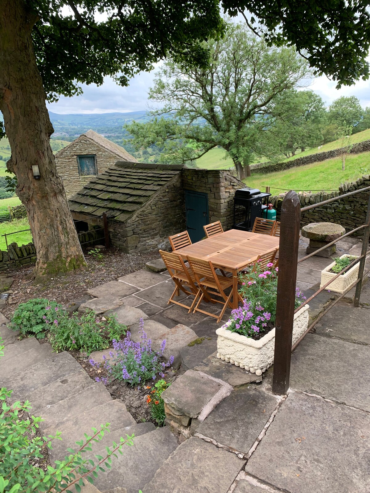 An outdoor terrace features a wooden dining table surrounded by several folding chairs. Stone pathways are lined with flower planters, while a small stone building with a green door is set against a backdrop of rolling hills and trees.