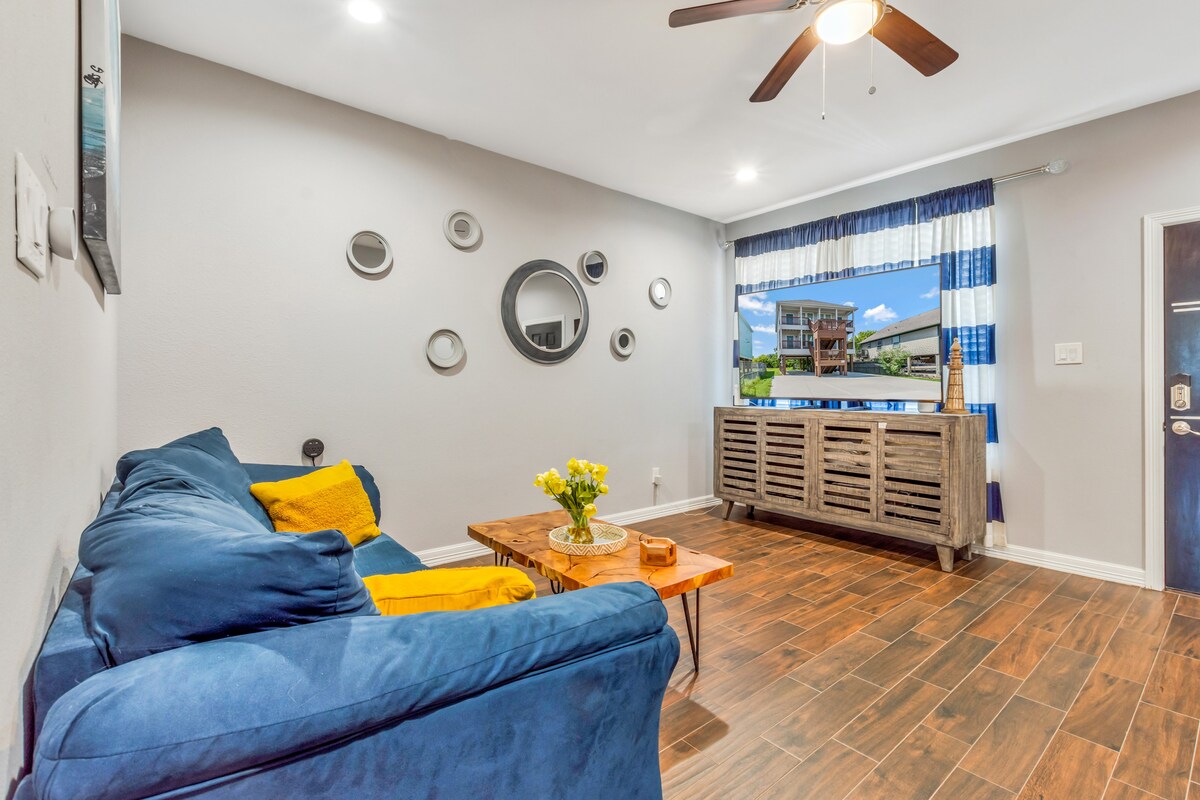 A comfortable living area features a blue couch adorned with a yellow throw pillow. A wooden coffee table sits in front, with a vase of flowers on top. Decorative mirrors are mounted on the wall beside a window dressed with blue and white curtains, allowing natural light to illuminate the space.
